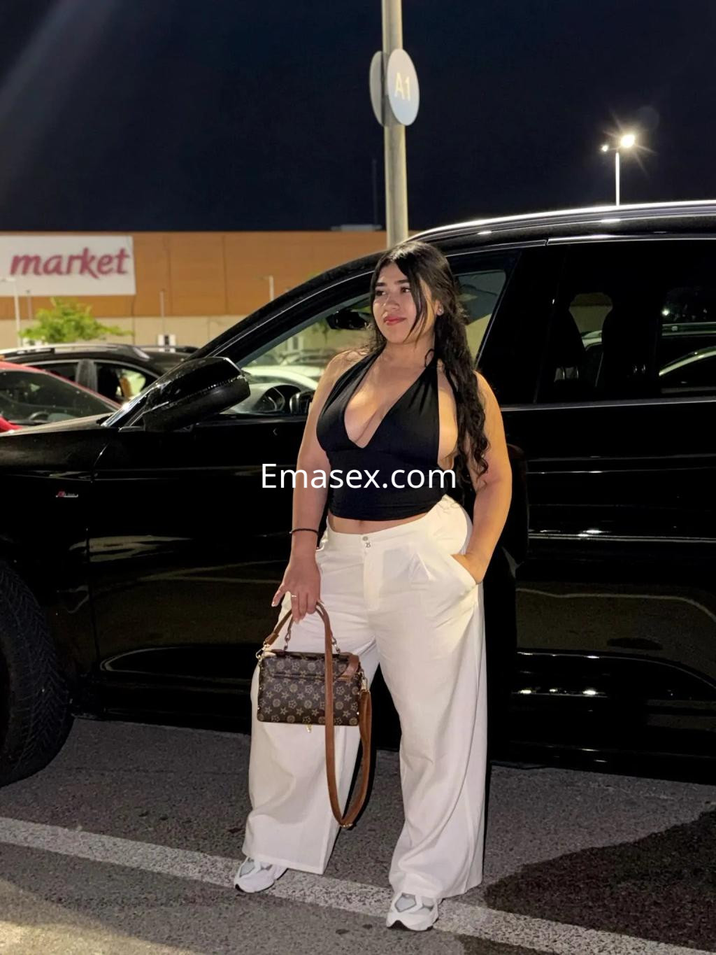 La puta posa junto a un coche negro, luciendo un top negro y pantalones blancos en Palma de Mallorca.