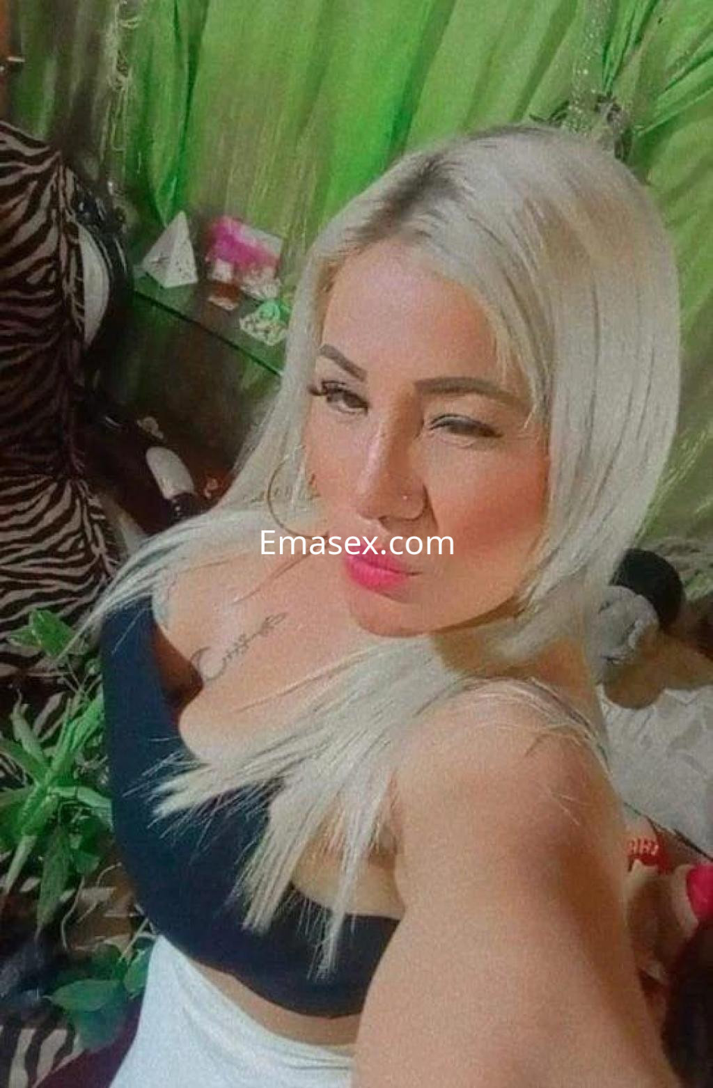 Escort en Barcelona, posando en un entorno con plantas, luciendo una blusa negra y cabello rubio largo.