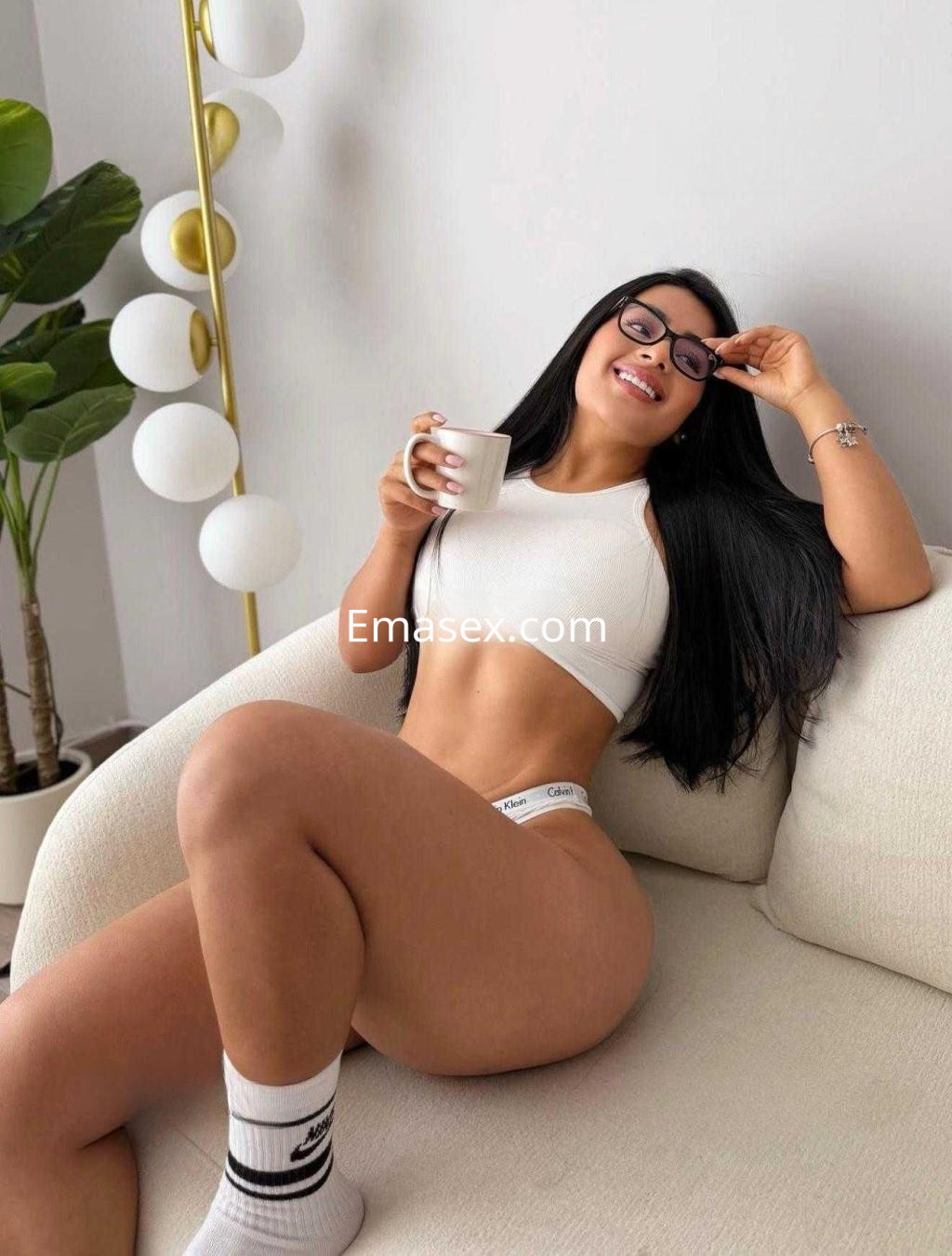 Escort en Barcelona con top blanco, jeans cortos y gafas, sentada en un sofá sosteniendo una taza.