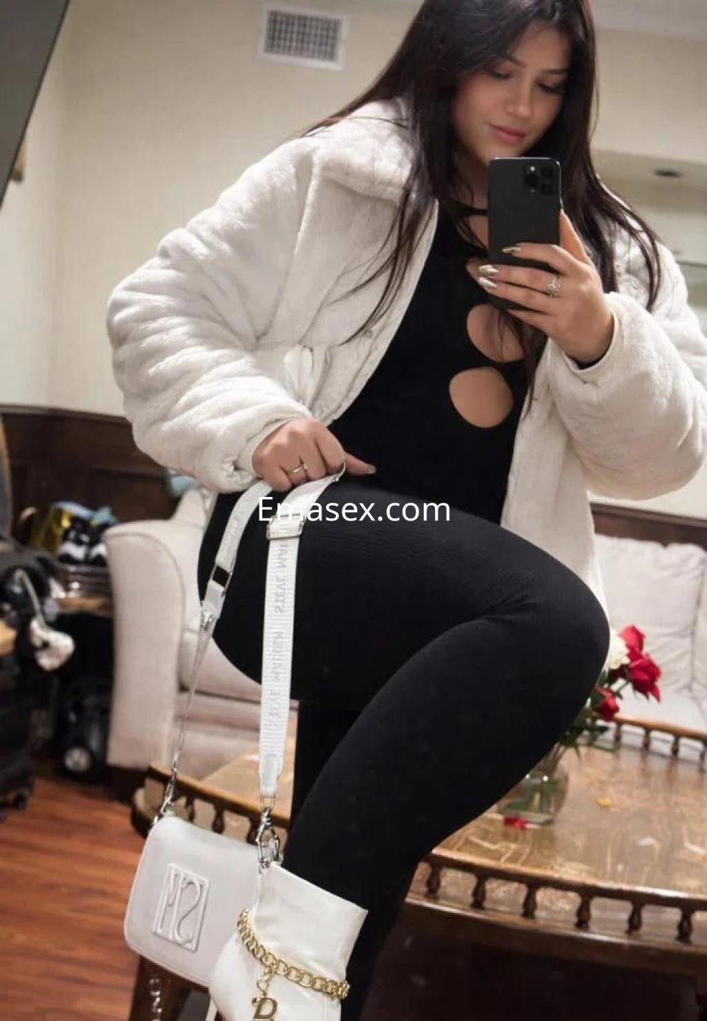 La puta en Lleida viste una chaqueta blanca y un conjunto negro, posando con una bolsa blanca en la mano.
