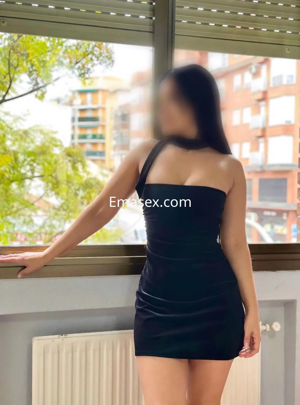 Escort en Madrid con vestido negro ajustado, posando junto a una ventana con vista a edificios.
