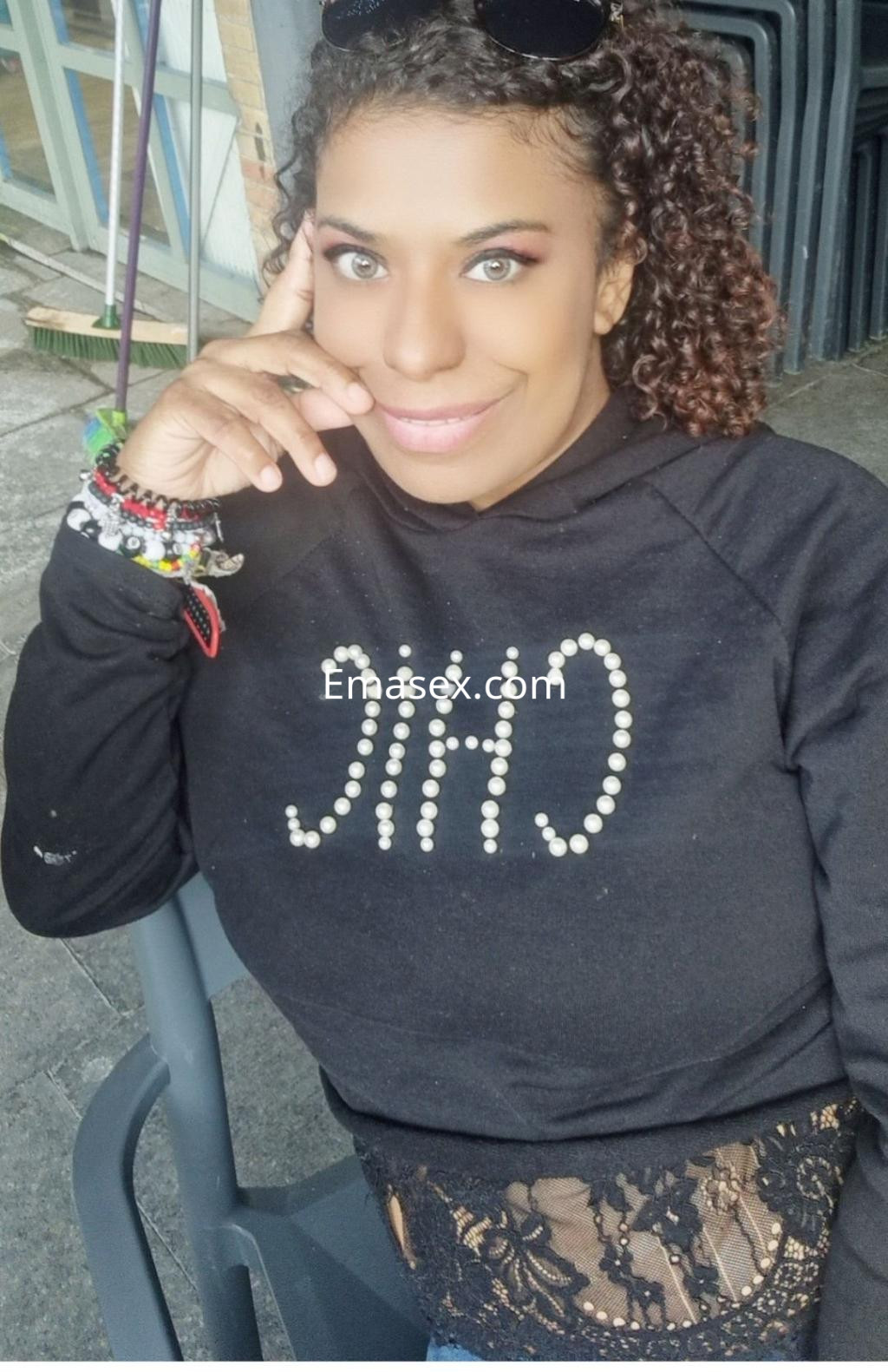 Escort en Málaga sonríe mientras se apoya en una silla, vistiendo una sudadera negra y con cabello rizado.