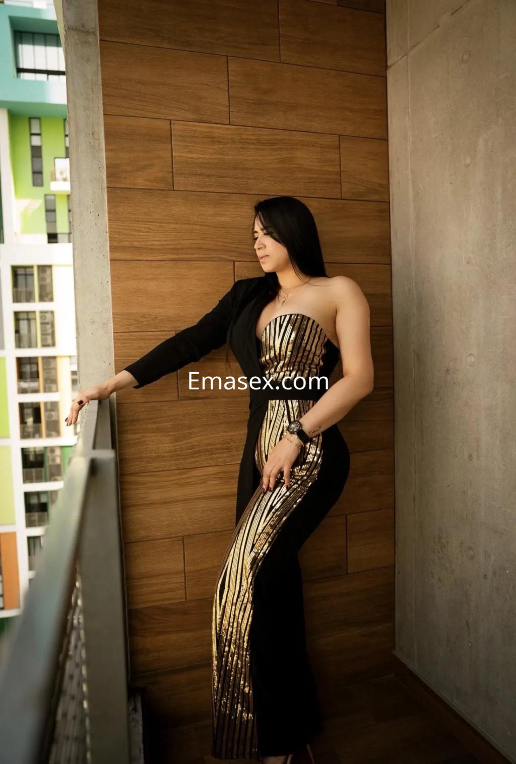La escort en Murcia posa de pie en un balcón, luciendo un elegante mono negro con detalles dorados.