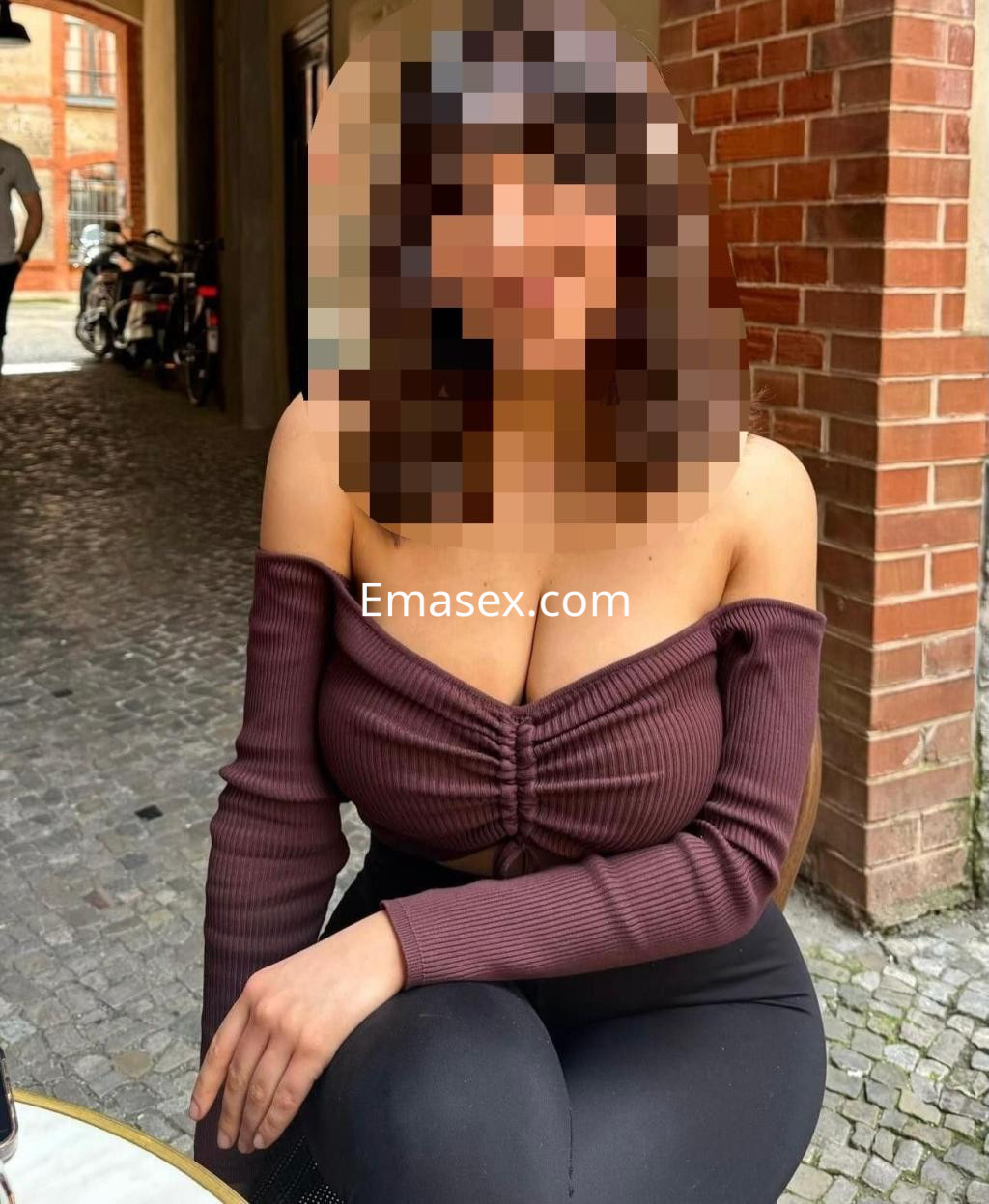 Puta sentada en una terraza en Murcia, con blusa de manga larga marrón y leggings ajustados, luciendo un peinado oscuro.