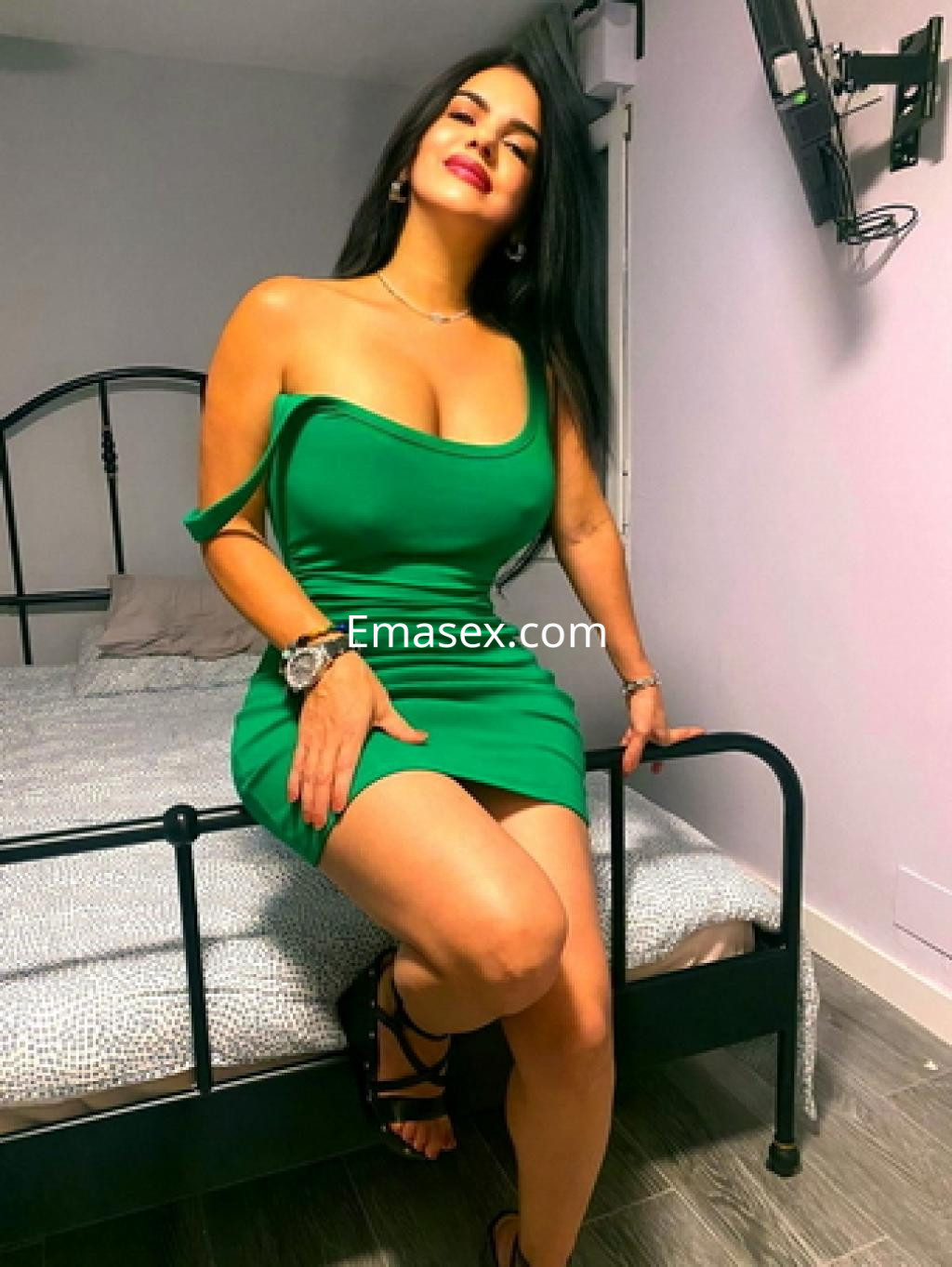 Escort en Salamanca luciendo un vestido verde ajustado, sentada en una cama con un fondo neutro.