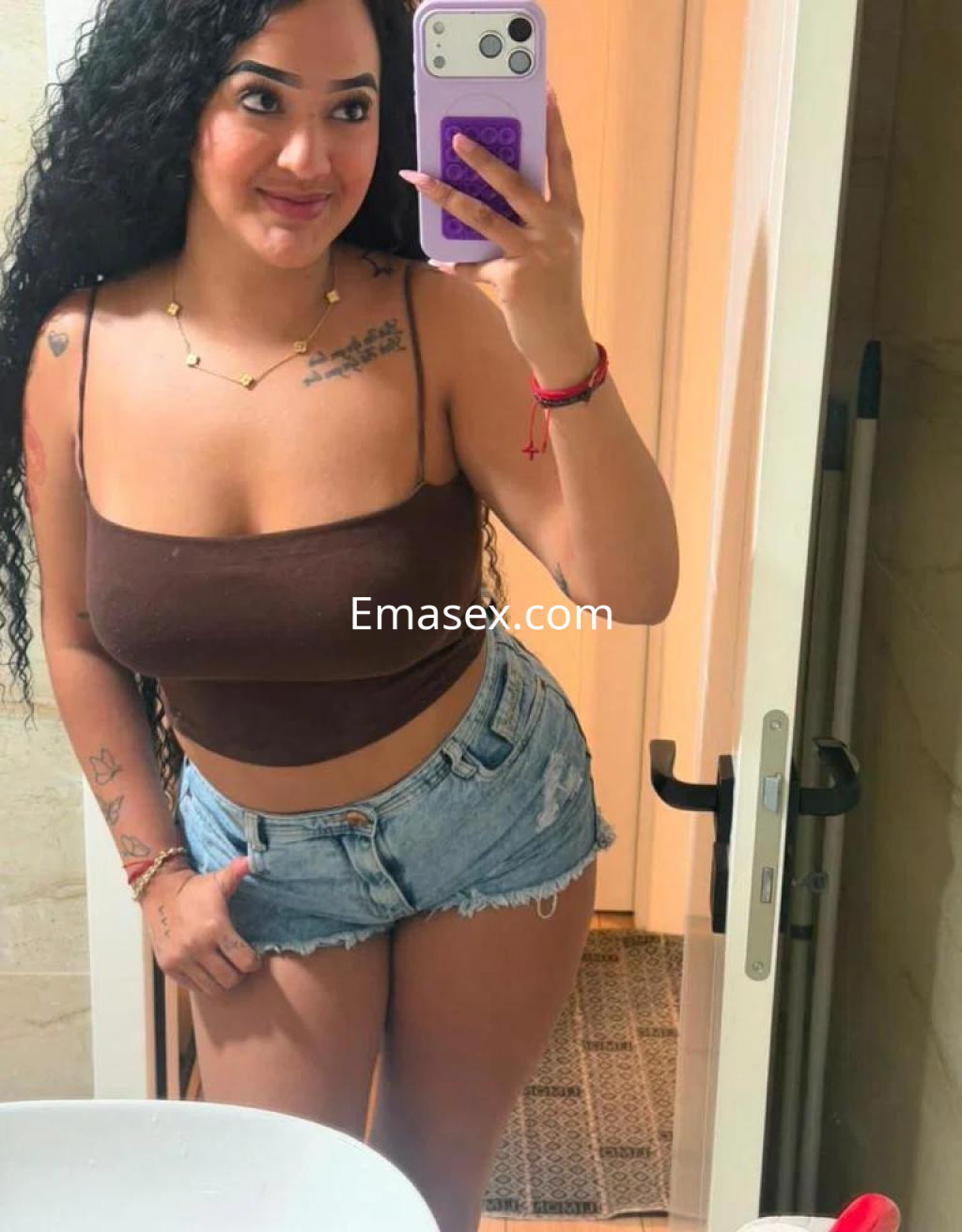 Escort en Santander, sonriendo frente al espejo con un top marrón y shorts de mezclilla, en un ambiente interior.