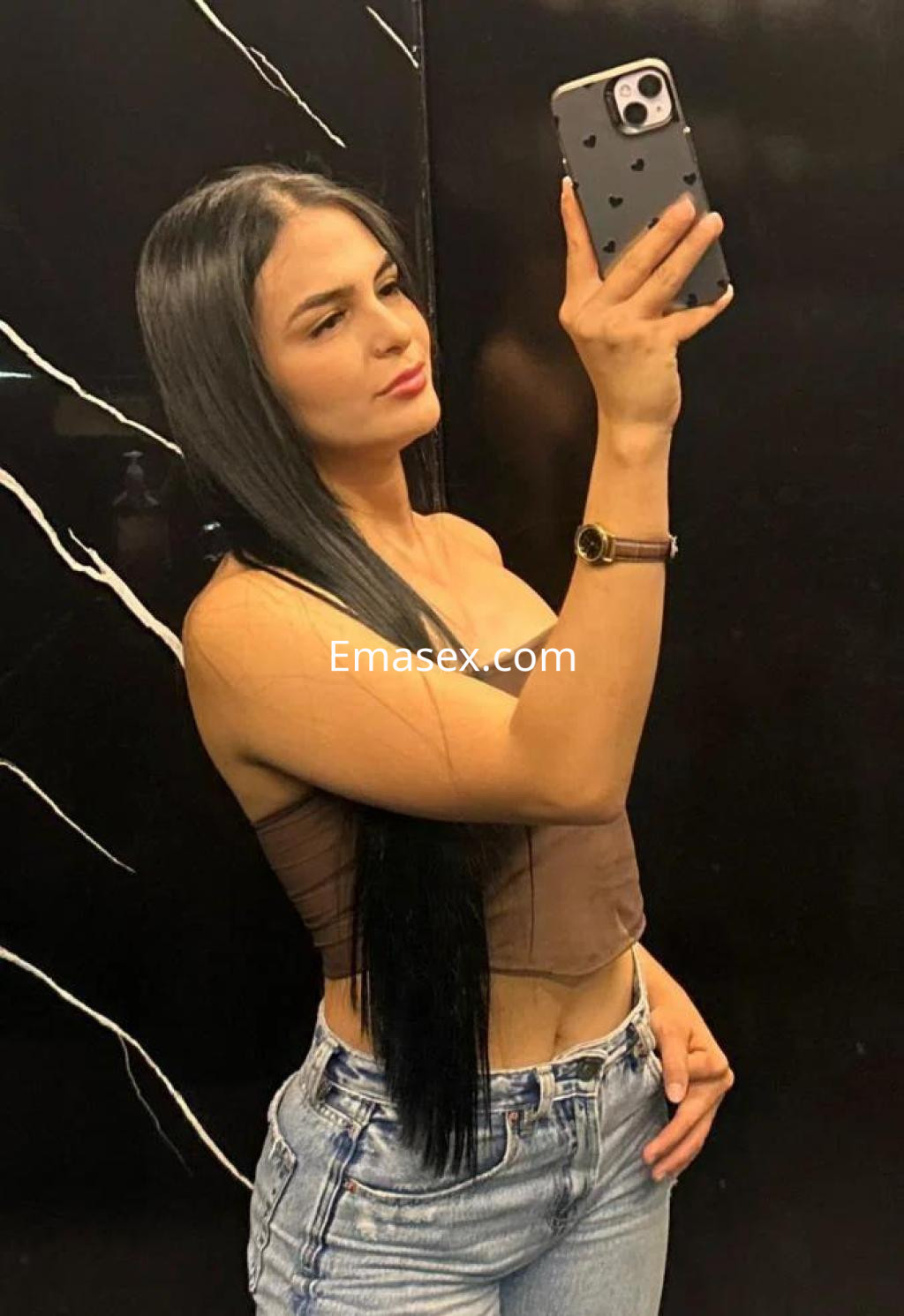 Escort en Tarragona con cabello largo y lacio, usando un top marrón y jeans ajustados, posando frente a un espejo.