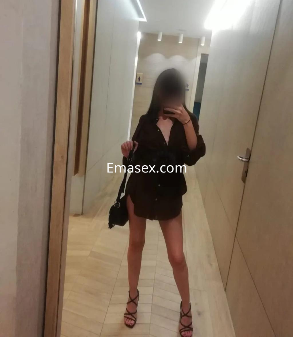 La puta posa en un pasillo con una camisa oscura, sandalias y una bolsa, mostrando sus largas piernas en Valencia.