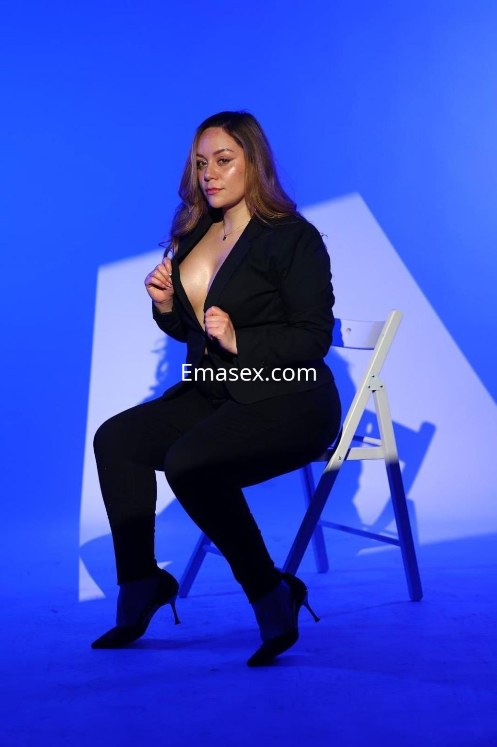 La puta en Valencia posa sentada en una silla blanca, vistiendo un traje negro con escote y tacones altos.
