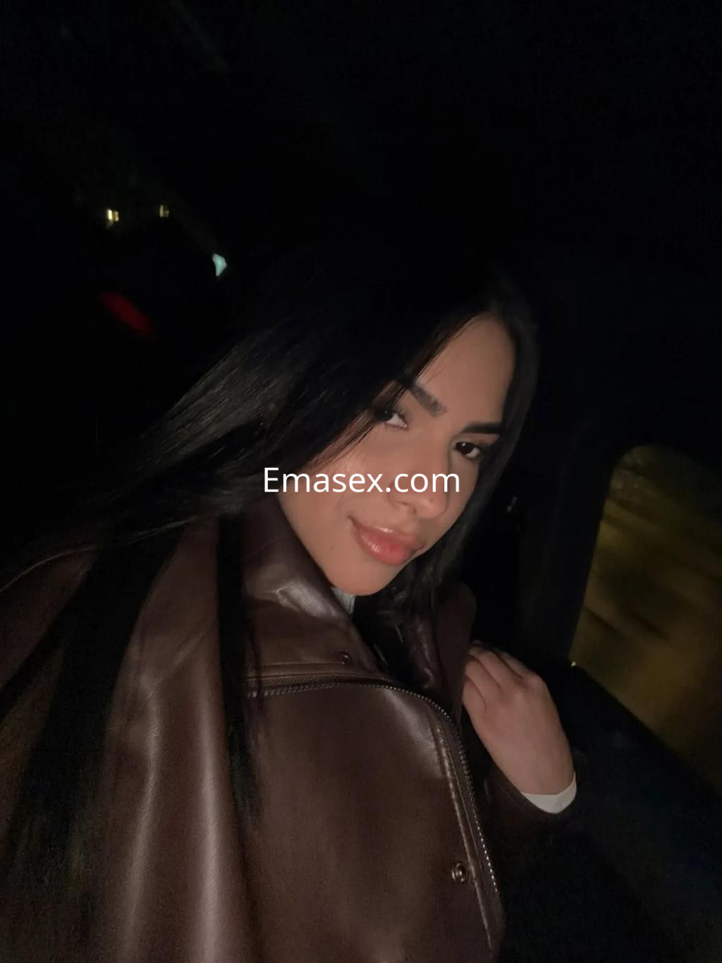 Puta con cabello largo y oscuro, viste una chaqueta marrón, posando en un ambiente oscuro en Valencia.