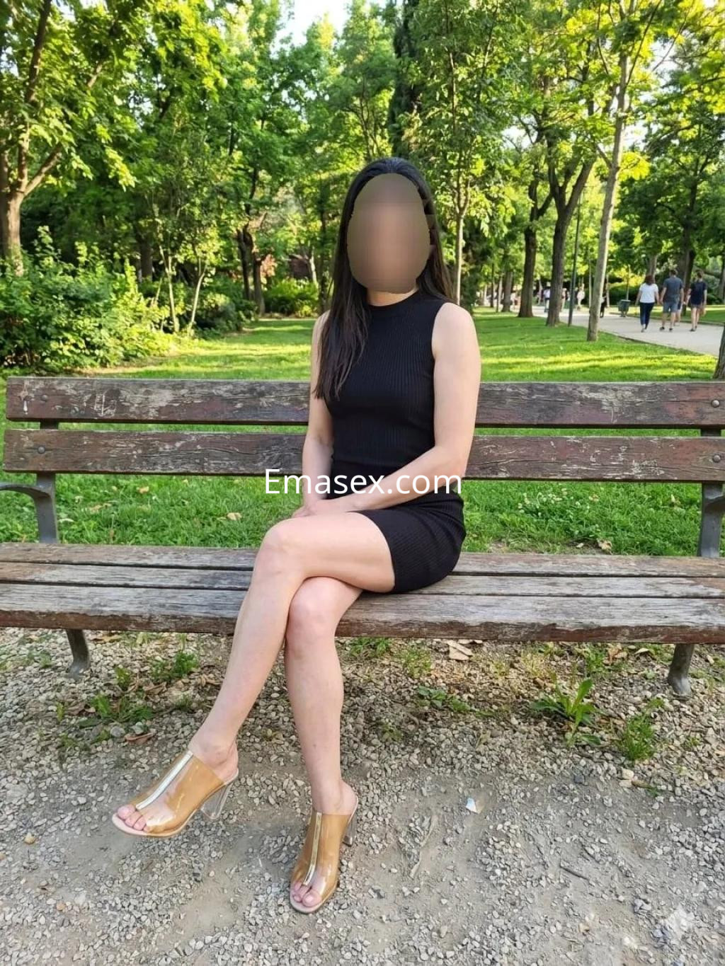 Puta sentada en un banco de parque en Valencia, vistiendo un vestido negro ajustado y sandalias transparentes.