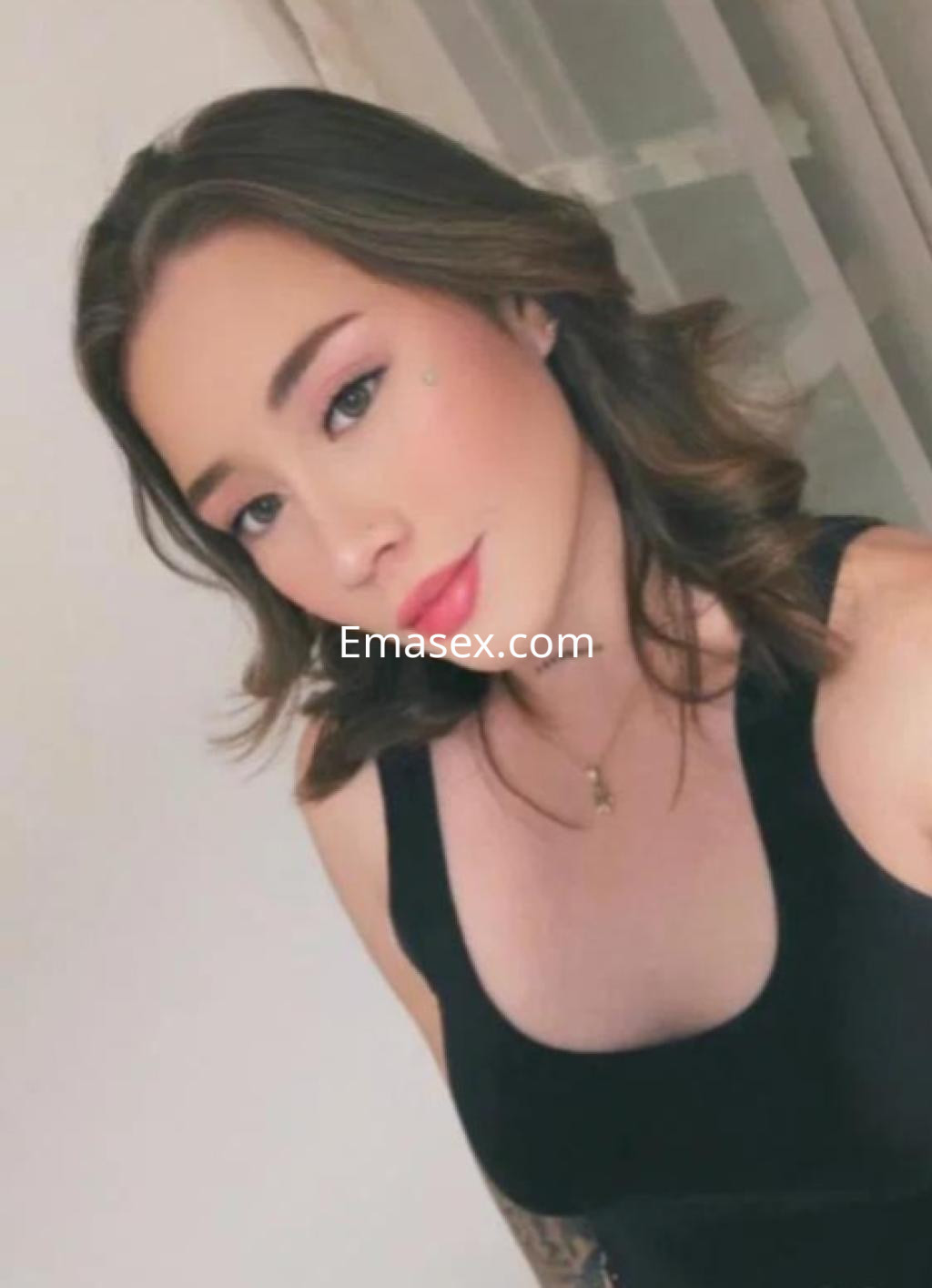 Puta en Gijón con cabello ondulado, blusa negra ajustada y maquillaje suave, posa con una expresión coqueta.