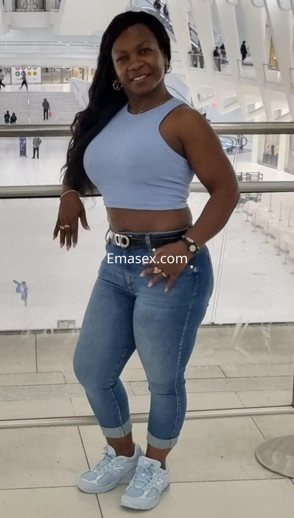 La puta en Málaga lleva una camiseta sin mangas azul y jeans ajustados, sonriendo mientras posa en un entorno moderno.