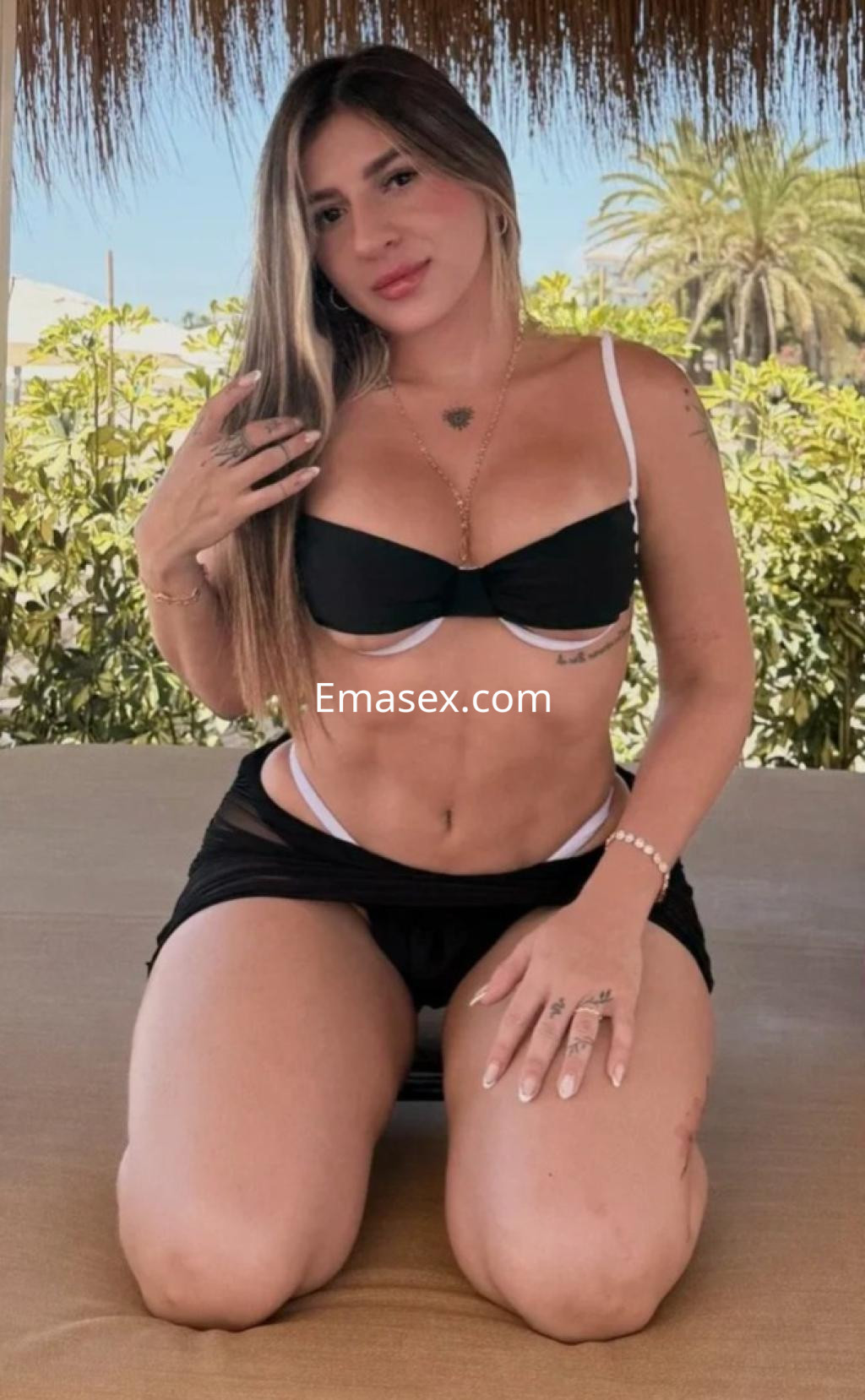 Puta en Málaga posando de rodillas con un top negro y shorts, mostrando su figura en un entorno exterior.