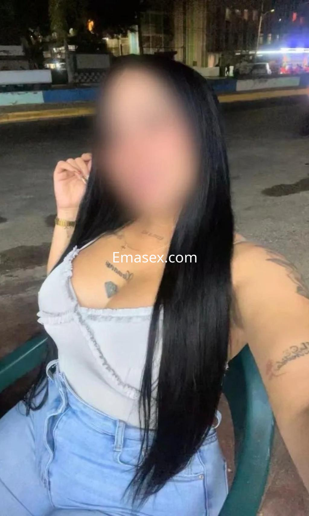 Escort en Valladolid con cabello largo y oscuro, vistiendo blusa clara y jeans, sentada en una silla al aire libre.
