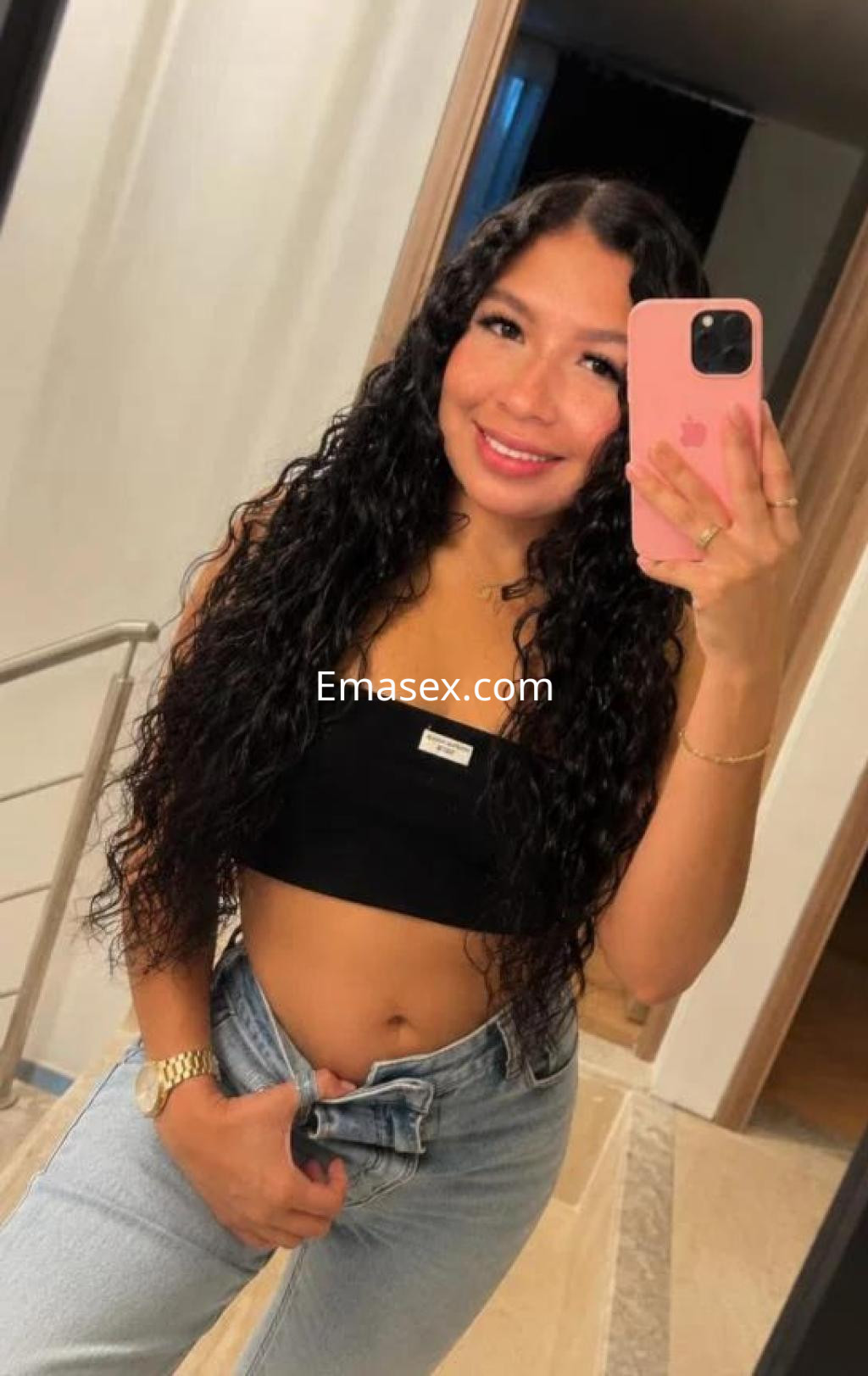Puta de Coruña se muestra sonriendo en un espejo, con blusa negra y jeans, cabello rizado y largo.