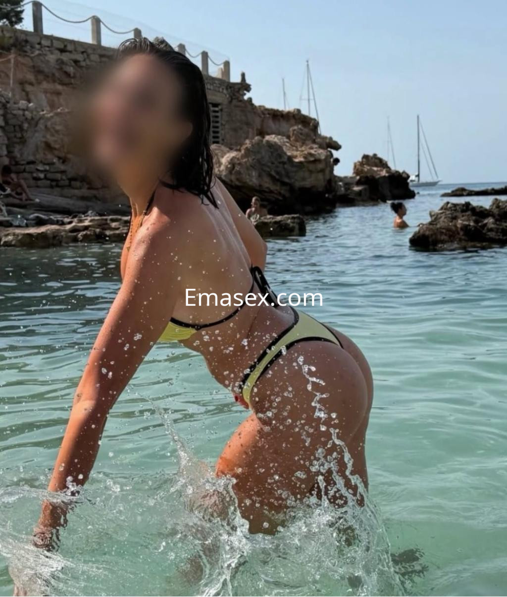 Escort en Irún, de cabello oscuro y húmedo, luciendo un bikini amarillo mientras juega en el agua.