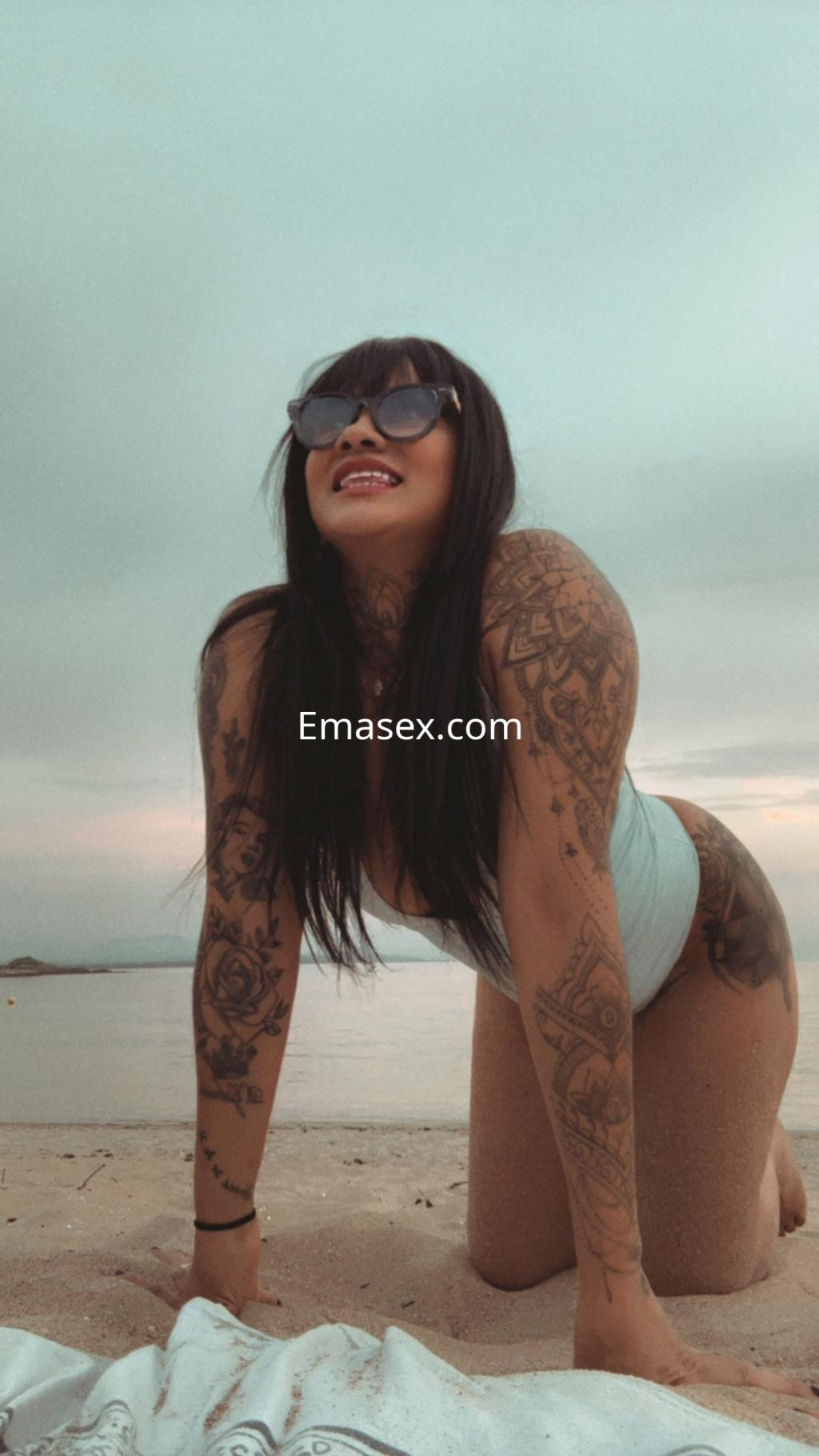 La puta de Ourense se encuentra en la playa, posando sobre las rodillas con un top claro y gafas de sol, mostrando sus tatuajes.