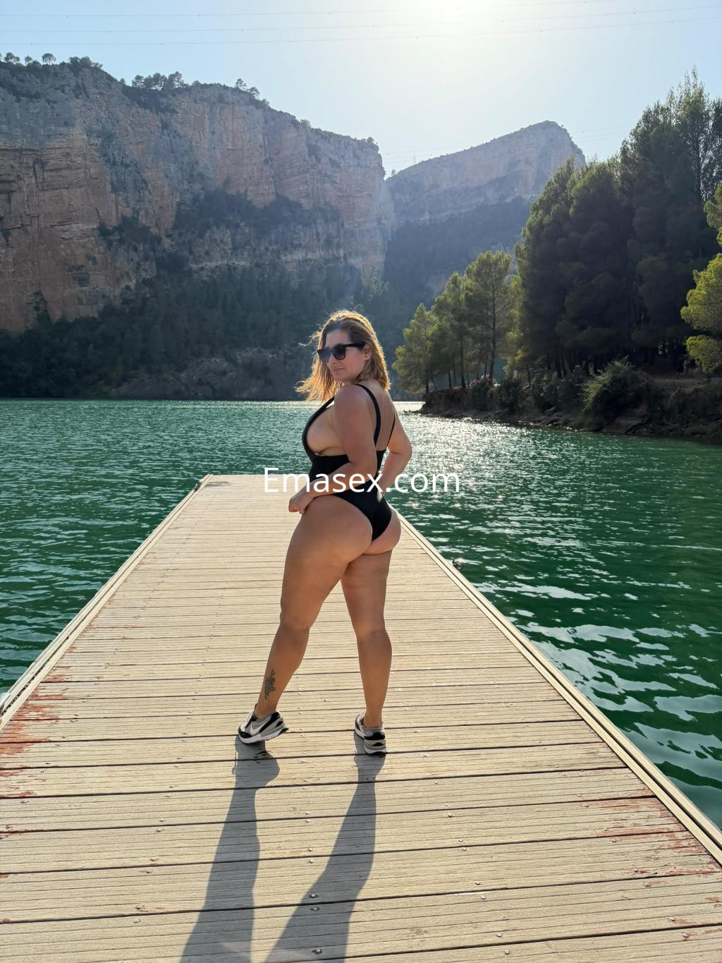 Escort en Santander luciendo un bañador negro, posando de perfil en un muelle junto a un lago rodeado de montañas.