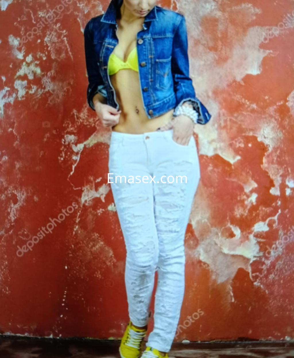 Escort en Vitoria viste un sujetador amarillo y jeans blancos ajustados, con chaqueta de mezclilla, en fondo rojo.