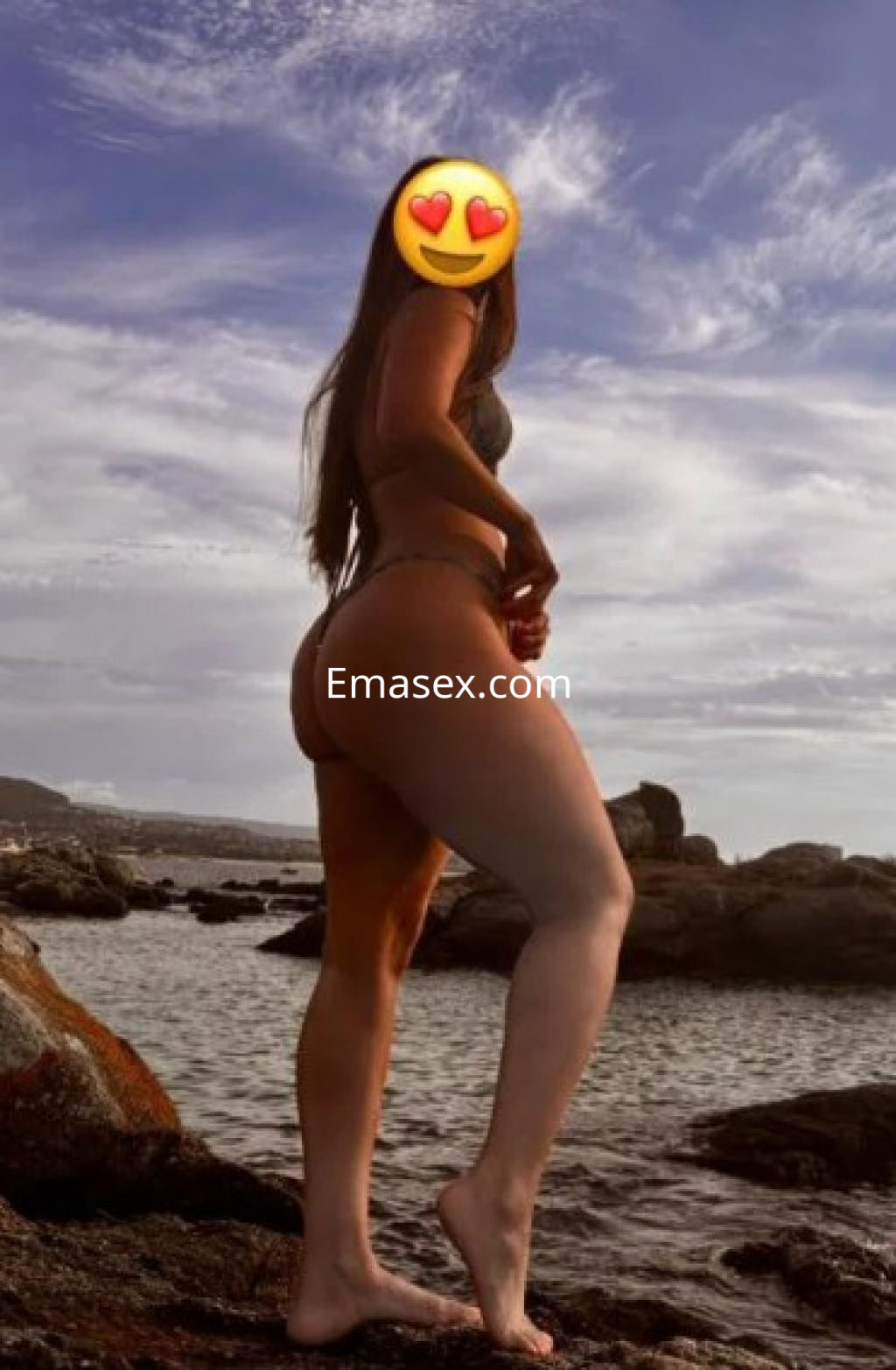 Escort en Vigo mostrando un bikini oscuro, con cabello largo y suelto, posando de perfil en la playa.