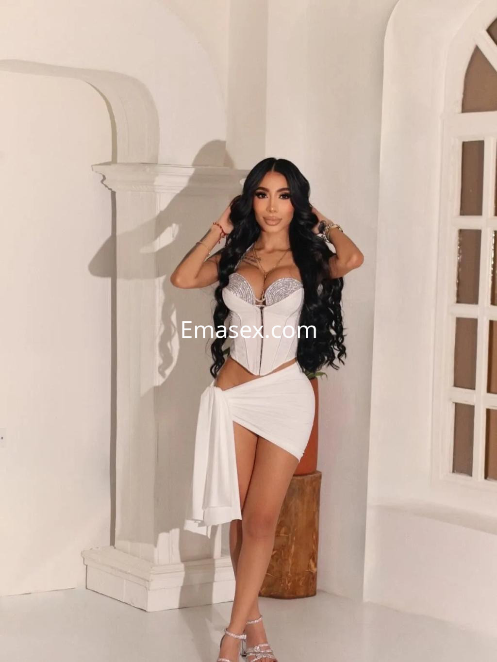 Escort en Alicante posando con un vestido blanco ajustado y detalles en lencería, con cabello largo y ondulado.