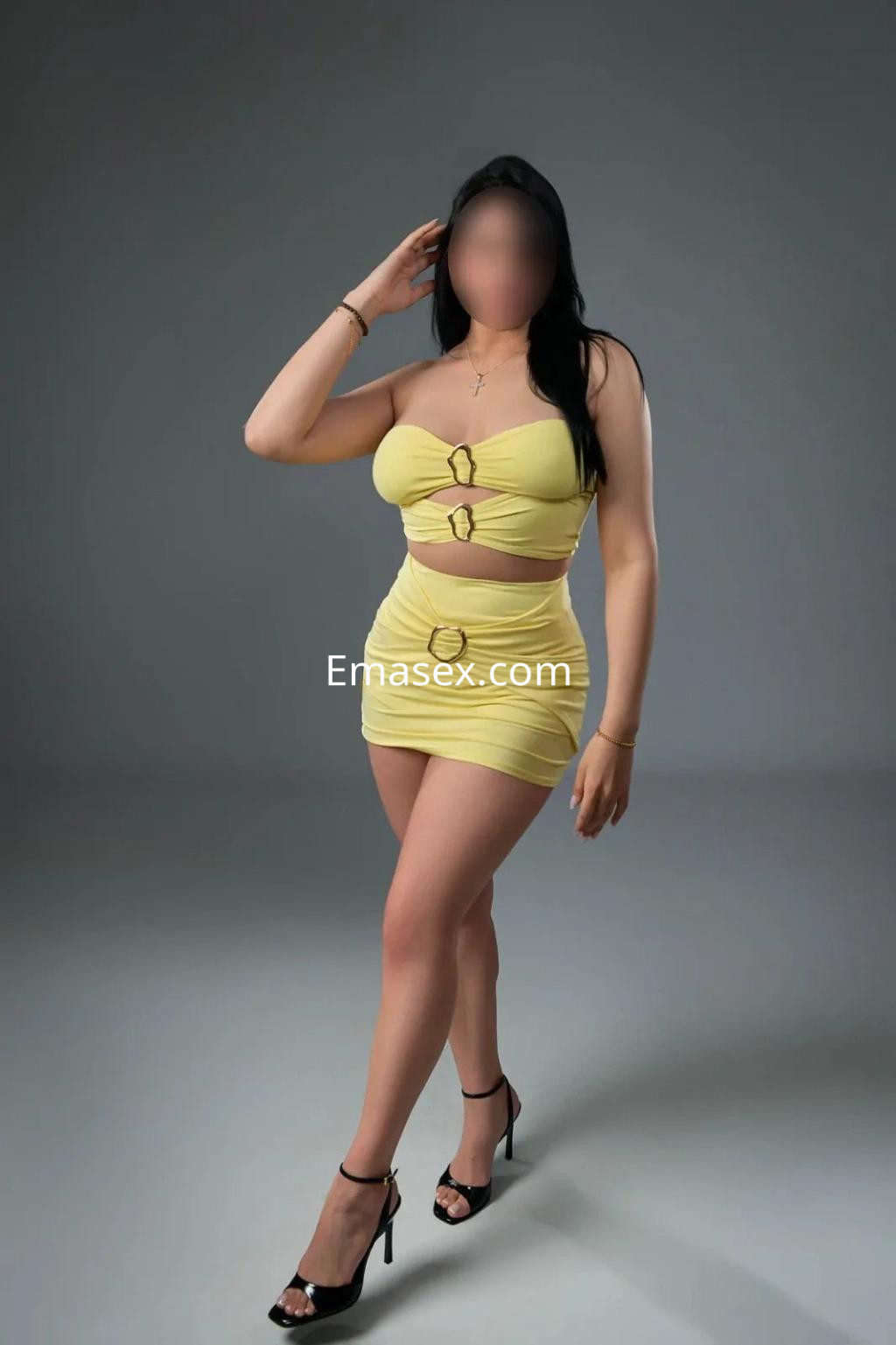 Escort en Alicante con cabello largo y oscuro, vistiendo un conjunto amarillo ajustado y tacones altos en un fondo gris.