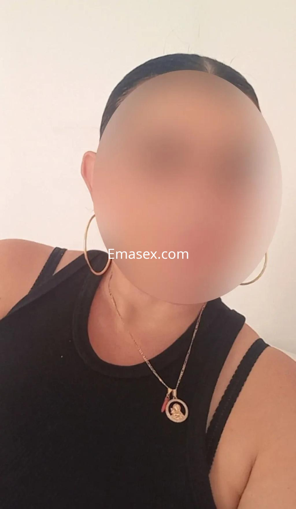 Puta con blusa negra sin mangas y aretes grandes, posando en un fondo claro en Alicante.