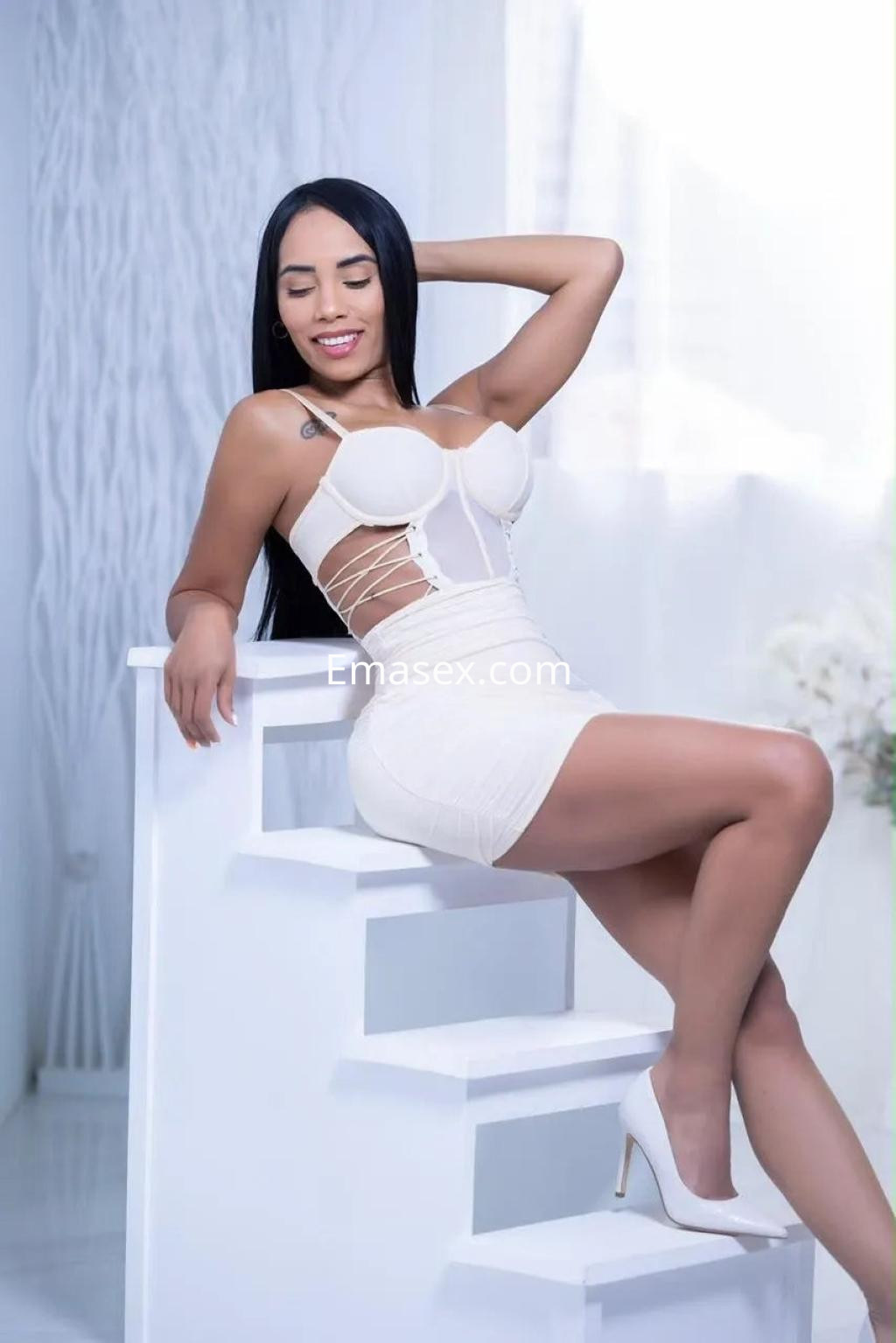 Escort en Alicante con cabello largo y lacio, viste un conjunto blanco y posa sentada en una escalera.