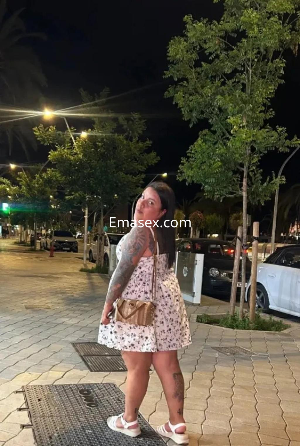 Escort en Palma de Mallorca con vestido blanco floral, posando sonriente en una calle iluminada por la noche.