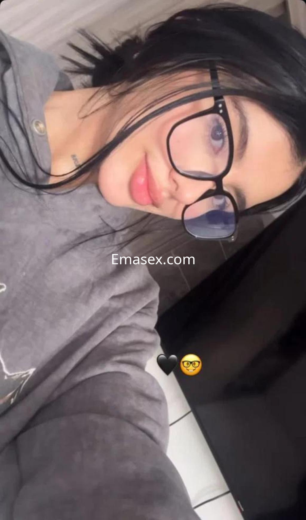 Puta en Huelva con cabello oscuro y lacio, usando gafas grandes y un sudadera gris en un ambiente interior.