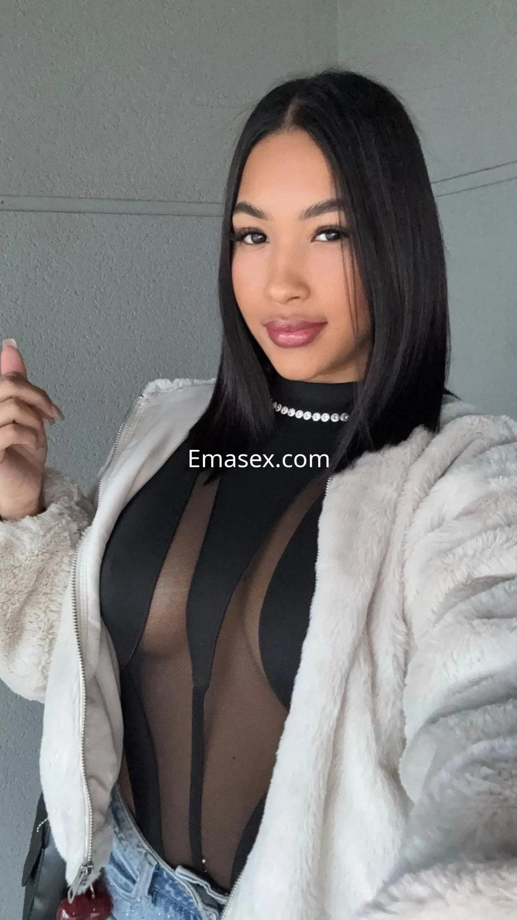 La puta en Murcia luce un body transparente con chaqueta de peluche y jeans, posando con una mano en el cabello.