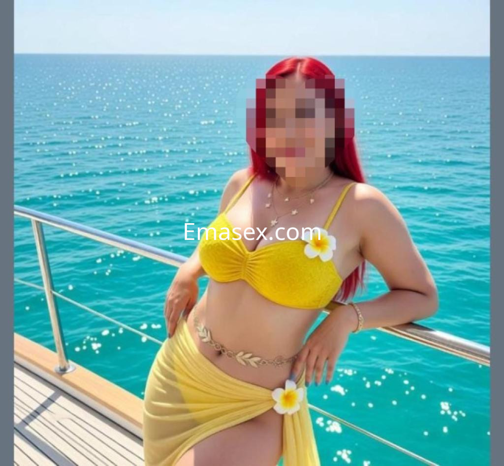 Escort en Ourense con cabello rojo, usando un bikini amarillo y una pareo, posando junto al mar.