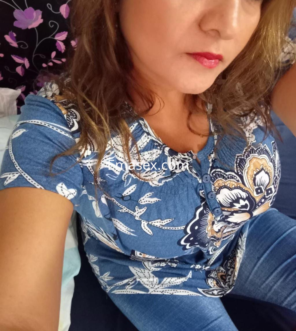 Puta en Toledo luce blusa azul con estampado floral y jeans, con cabello ondulado y labios rojos.