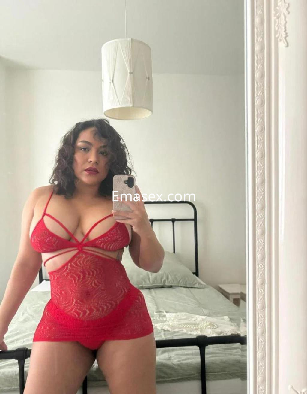 Escort en Toledo luciendo un conjunto de lencería roja con tiras, posando frente a un espejo en un dormitorio.