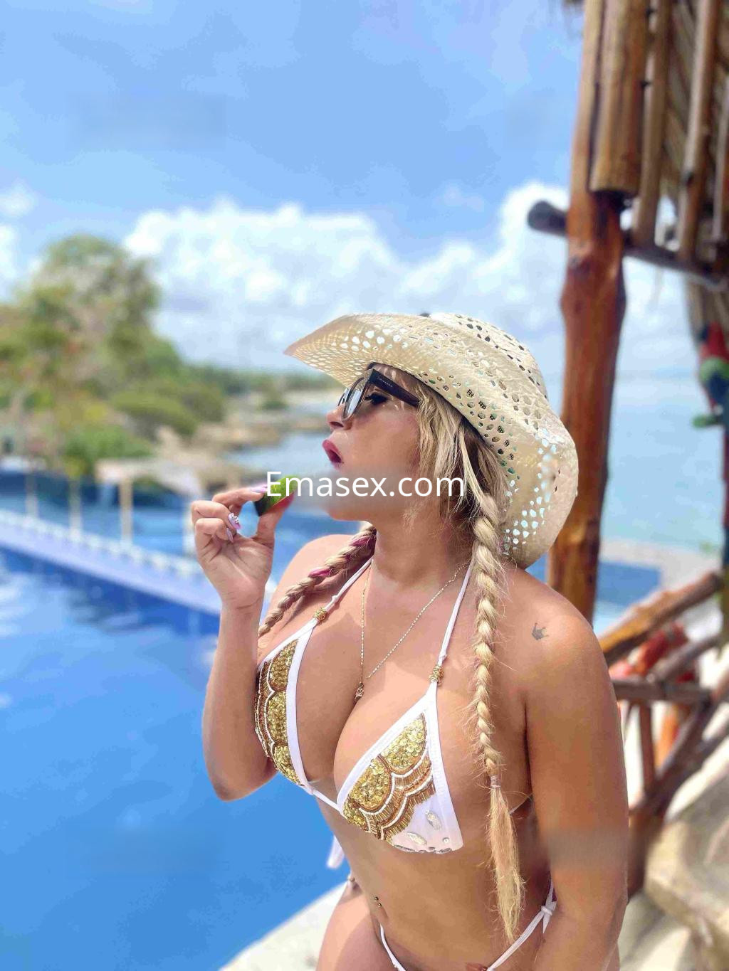 Escort en Zaragoza posando junto a una piscina, lleva un bikini blanco con detalles dorados y un sombrero de paja.
