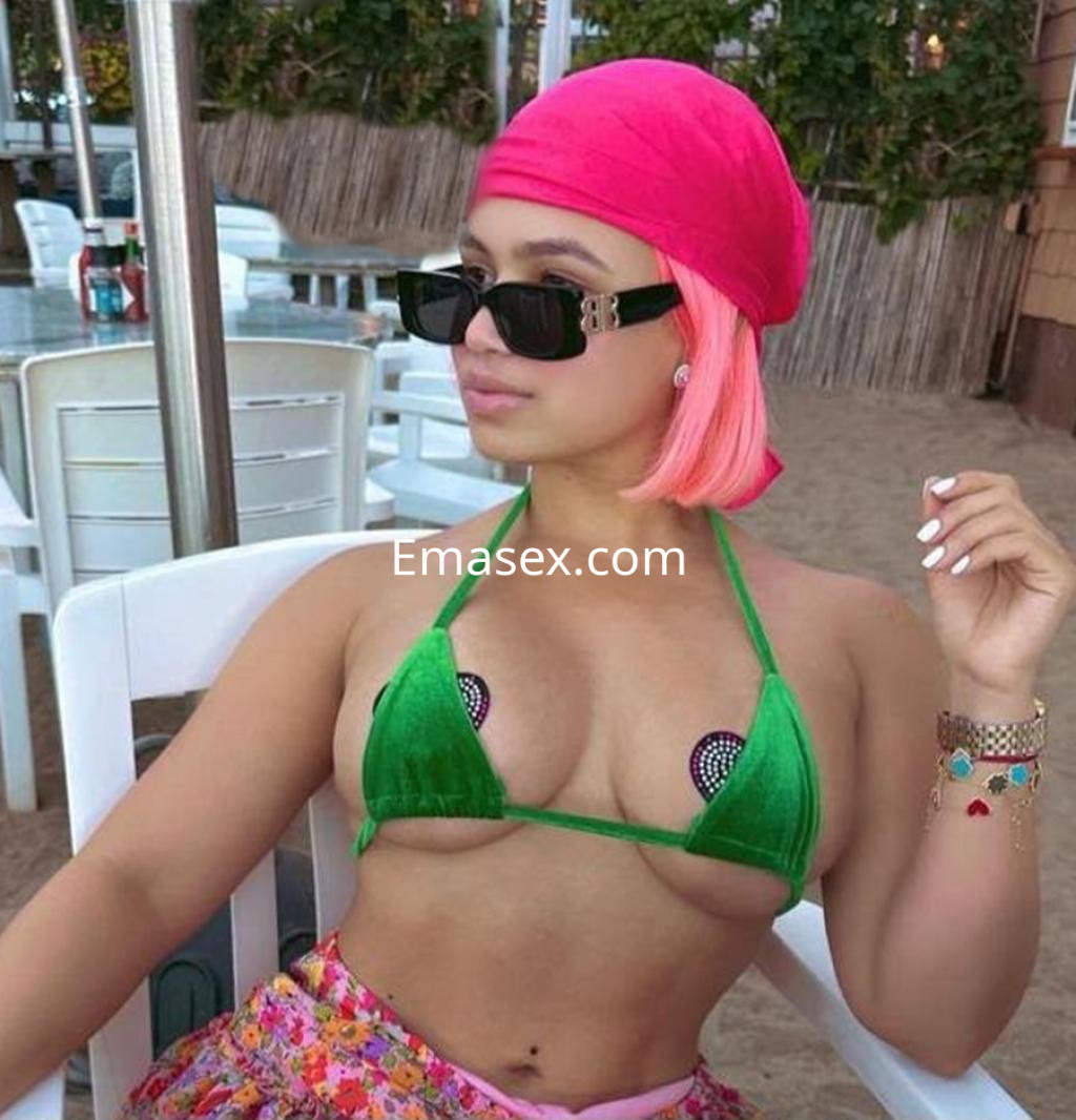 Escort en Pamplona con cabello rosa y bandana, vistiendo un bikini verde y una falda colorida, posando en la playa.