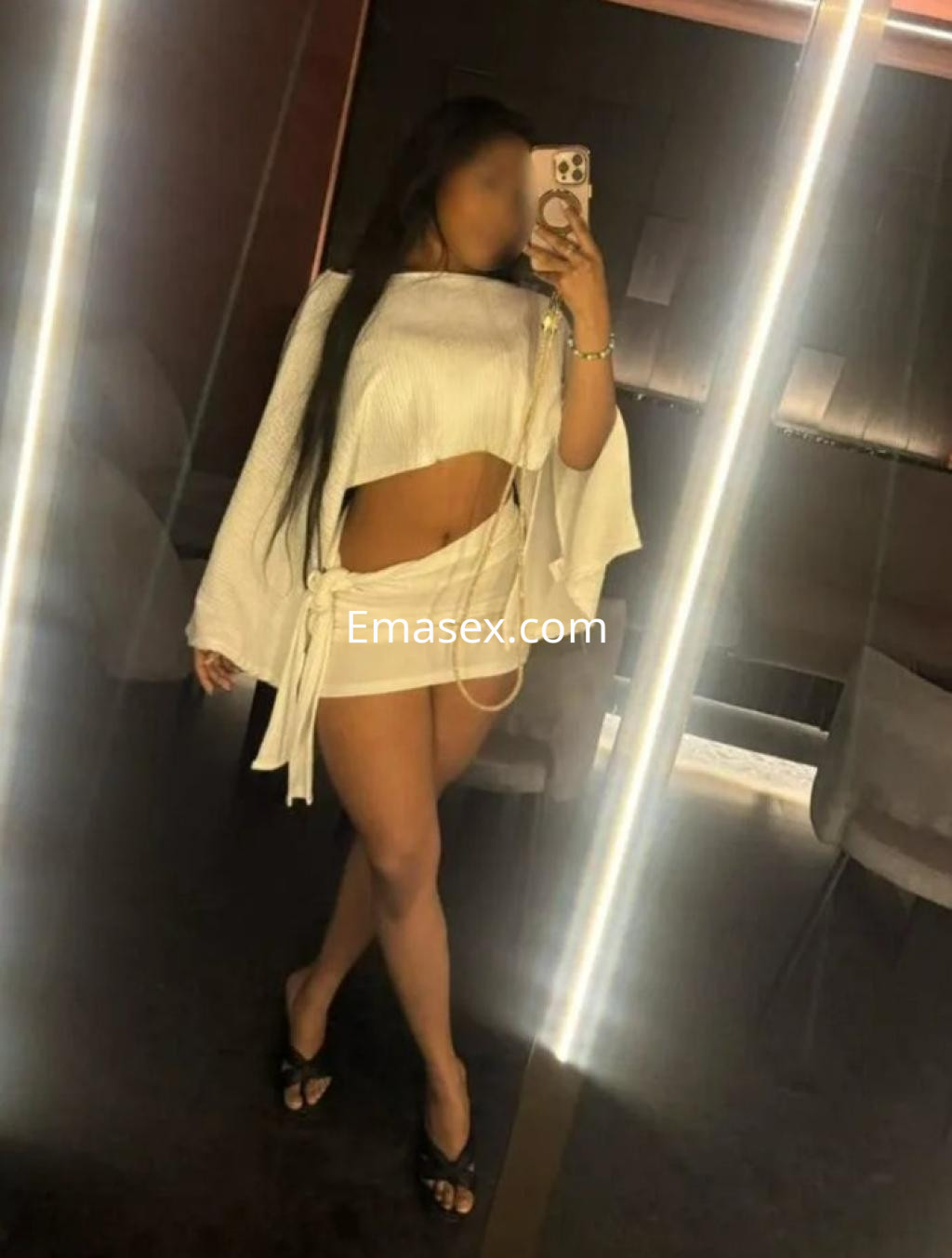 Puta en Madrid viste una blusa blanca con mangas amplias y una falda corta, posando frente a un espejo.