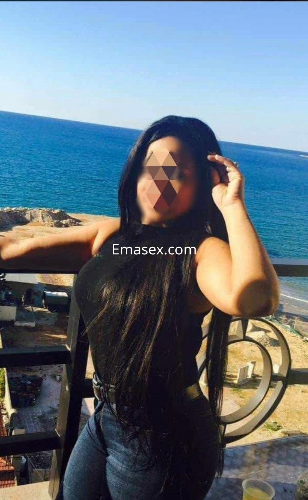 Escort en Vigo con cabello largo y oscuro, vistiendo una blusa sin mangas y jeans ajustados, posando en un balcón frente al mar.