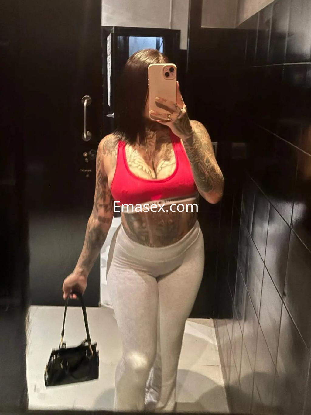 Escort en Barcelona posando en un espejo, viste un top rojo y pantalones blancos, con cabello corto y tatuajes visibles.
