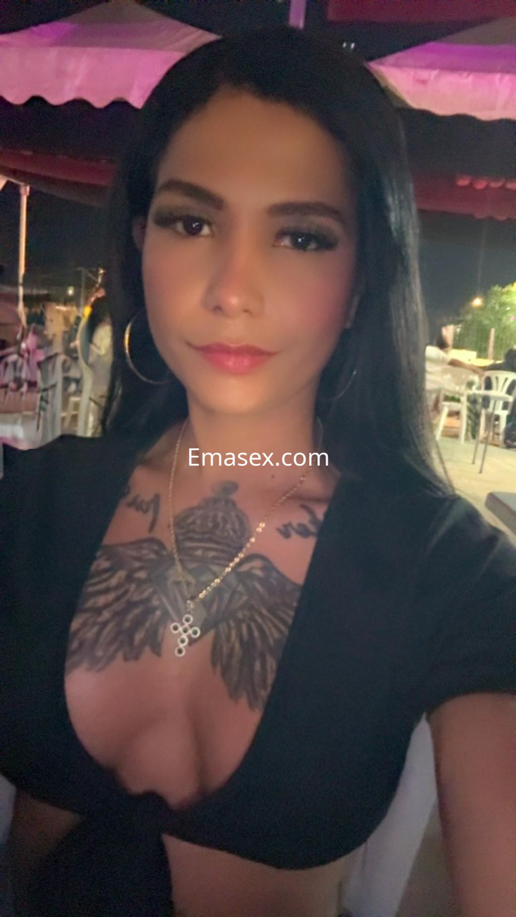 Puta trans en Valladolid, posando en un ambiente nocturno con una blusa negra y cabello largo, mostrando tatuajes en el pecho.