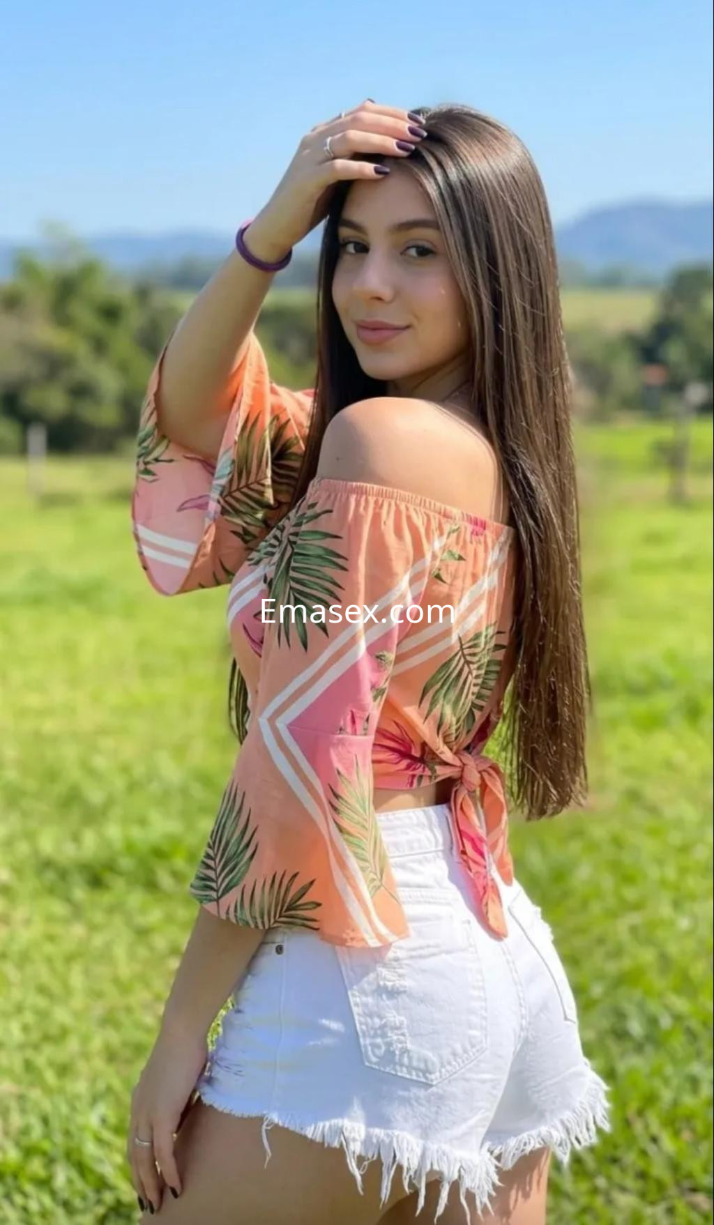 Escort en Murcia, vistiendo blusa de manga larga con estampado tropical y shorts blancos, posa al aire libre.