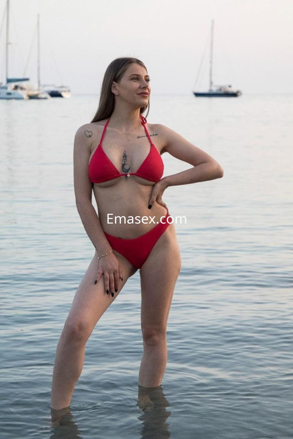 Escort en Palma de Mallorca, posando en la playa con un bikini rojo, mostrando tatuajes y el mar de fondo.