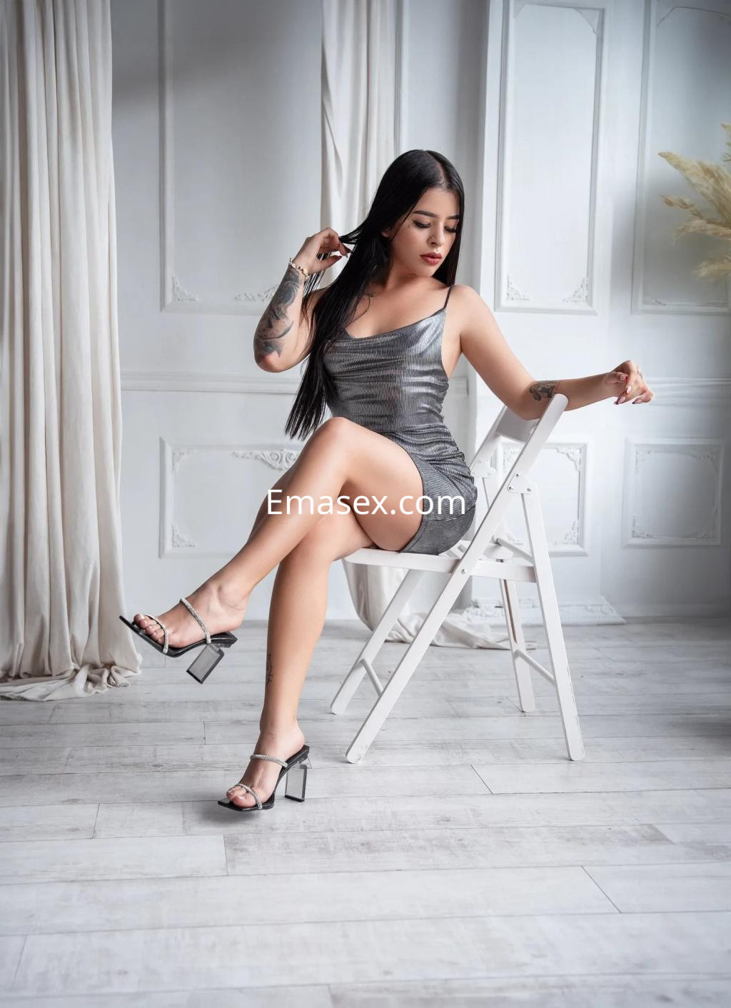 Escort en Palma de Mallorca luciendo un vestido plateado, sentada en una silla blanca, con cabello largo y lacio.
