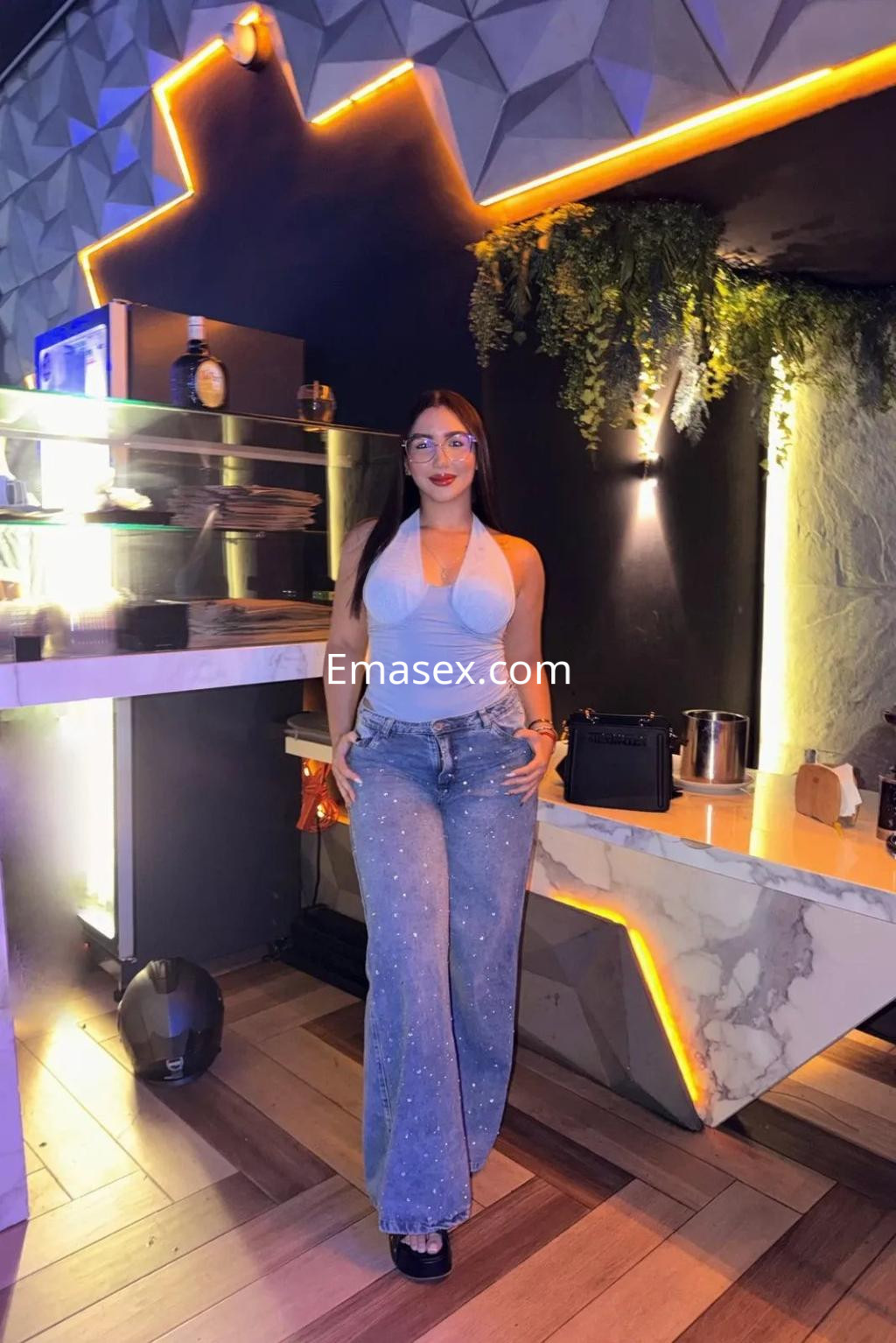 Puta de Barcelona con blusa gris y pantalones vaqueros decorados, posando en un ambiente moderno y luminoso.