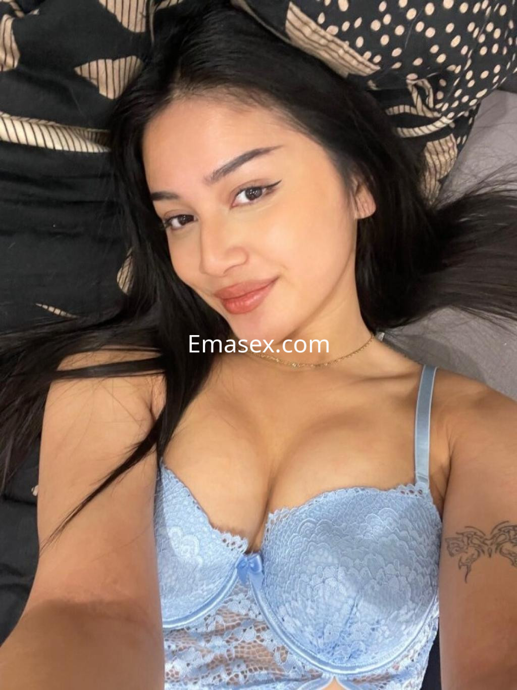 Escort en Burgos posando con una blusa de encaje azul, en un entorno de cama con sábanas de patrones oscuros.