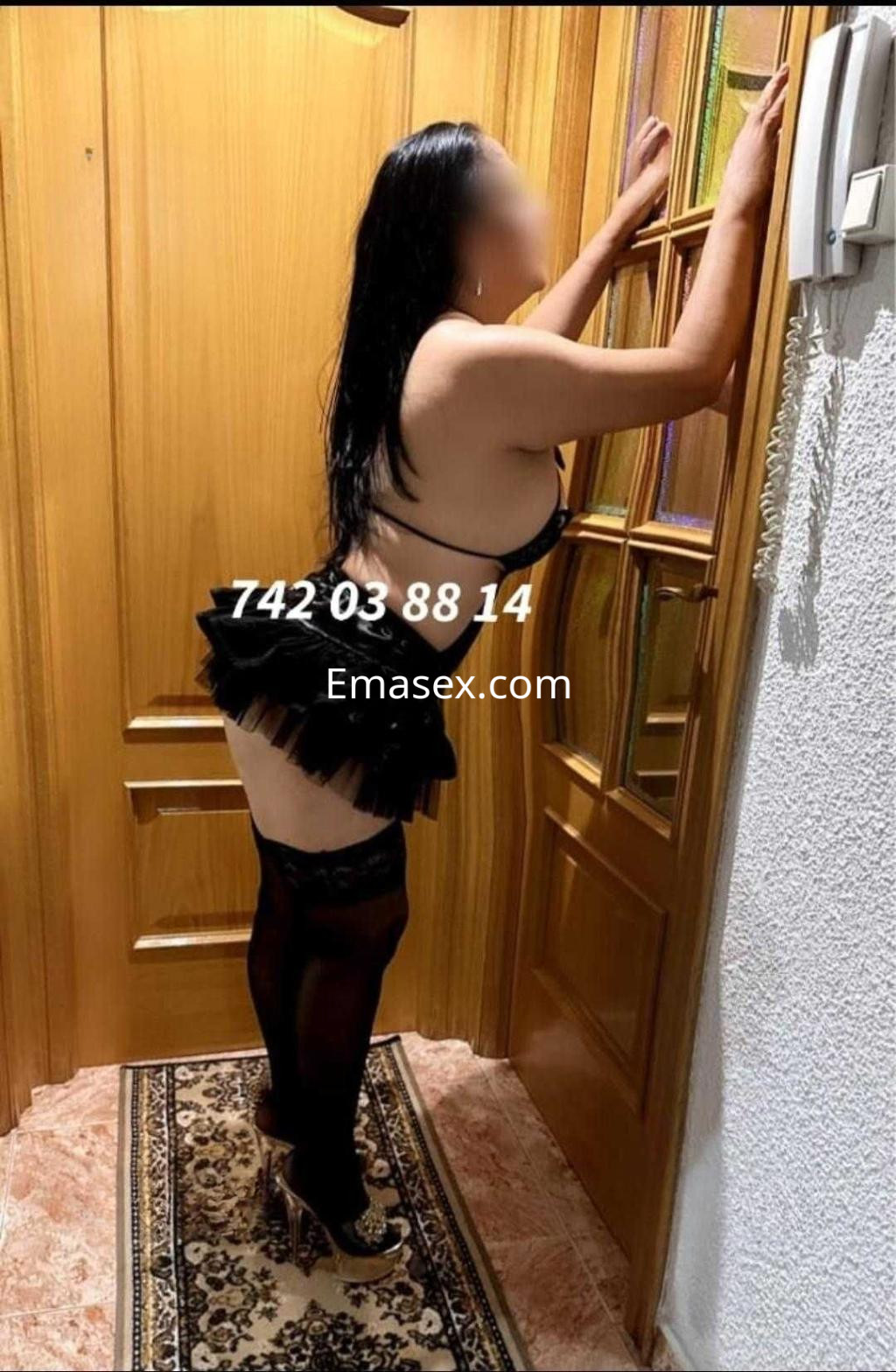 Puta en Cáceres, con lencería negra y falda corta, posando de espaldas junto a una puerta de madera.