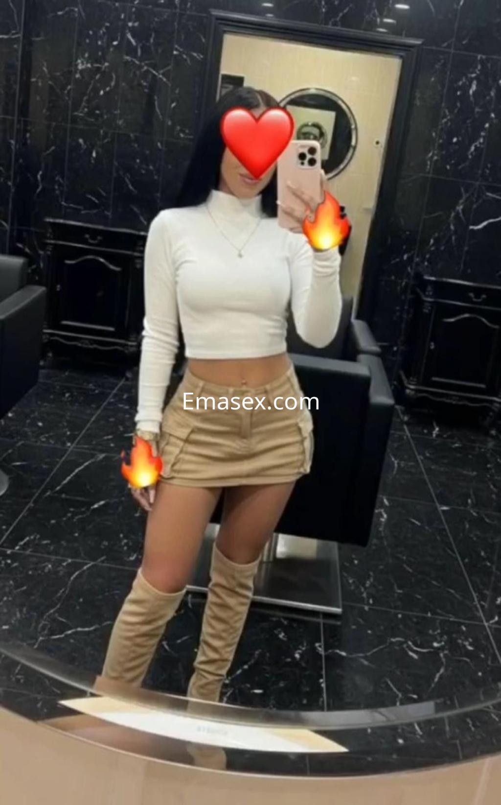 Puta en Coruña, con blusa blanca ajustada y falda de cuero, posando en un espejo con fondo oscuro.
