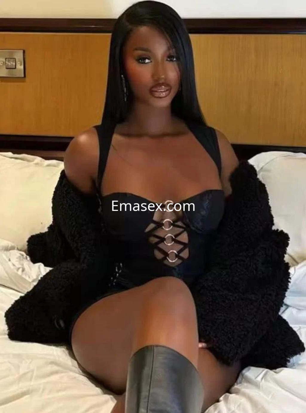 Escort en Girona luciendo un vestido negro ajustado y abrigo, sentada en la cama con una pose elegante.