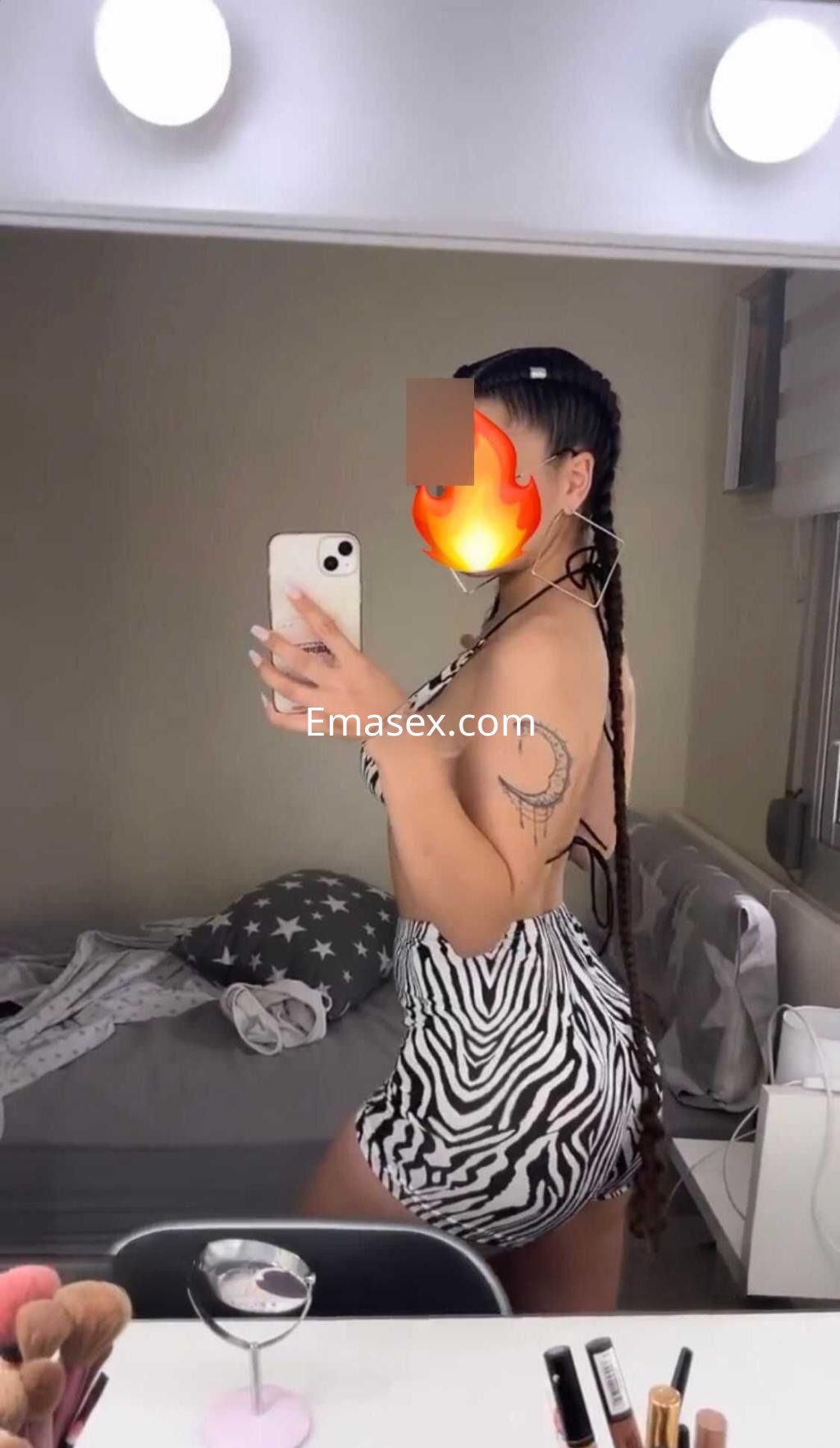 Puta en Huelva posando frente al espejo con un vestido de rayas y trenza larga, mostrando un tatuaje en el brazo.